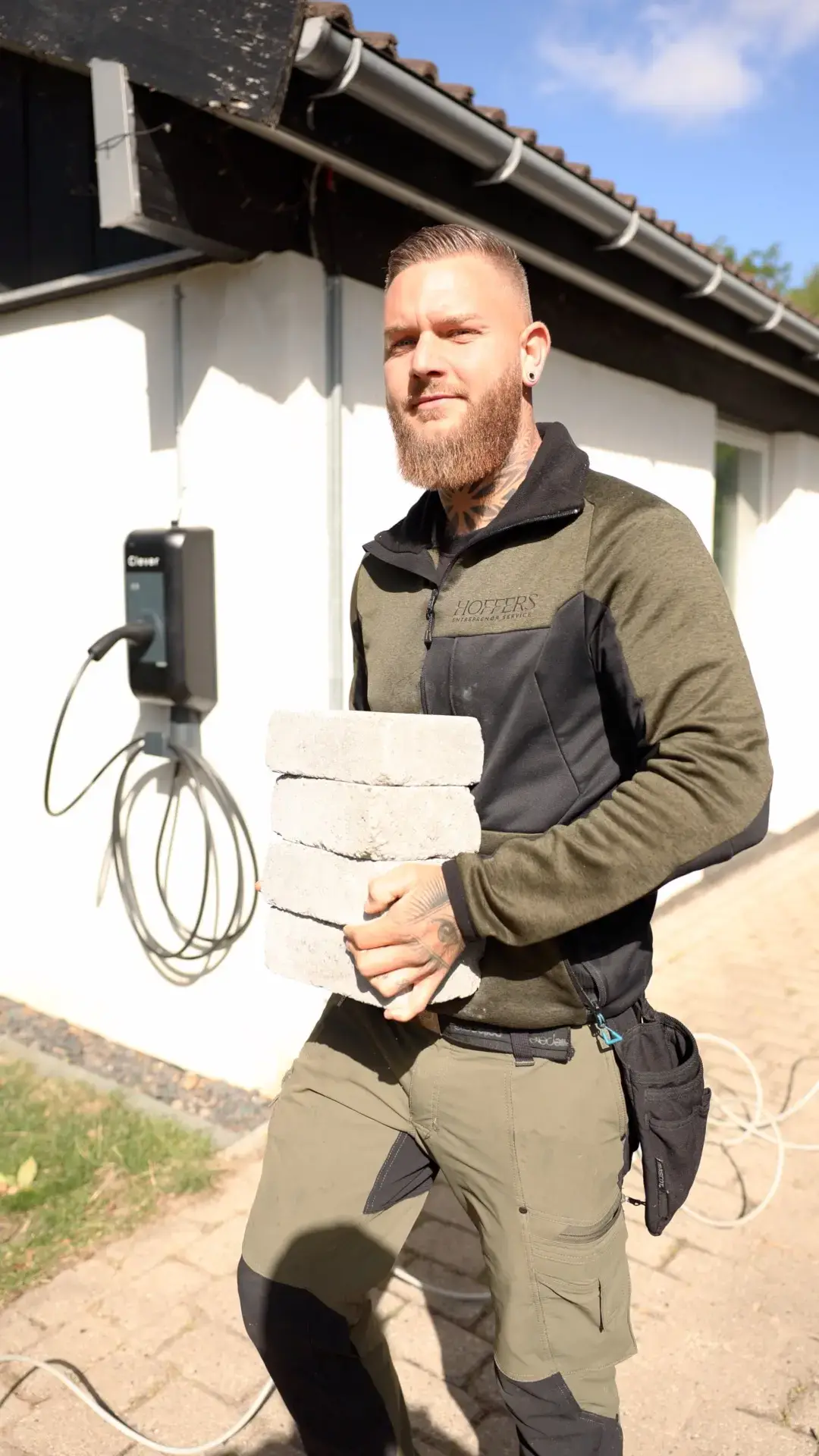 Mand med brunt hår og skæg holder mursten udenfor et hus med solskin.