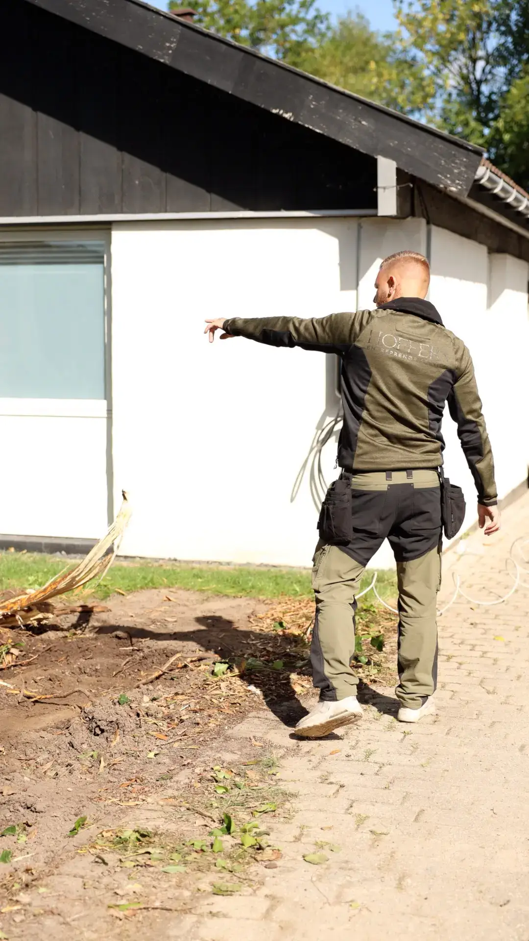 Mand peger mod væg udendørs foran et hus med sort tag og vindue.