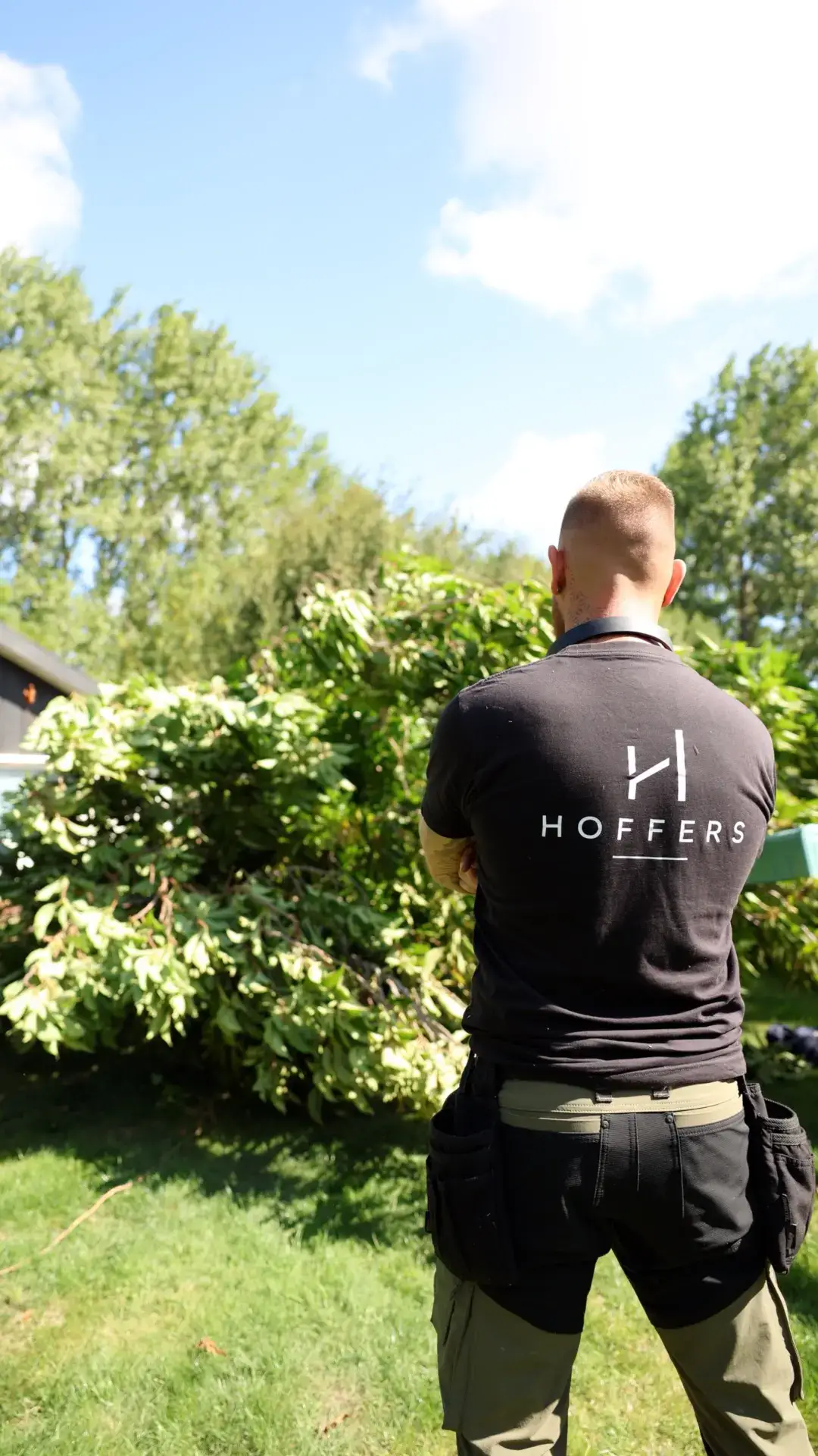Hoffers medarbejder i sort T-shirt med logo, står i grøn have med busk og træer.